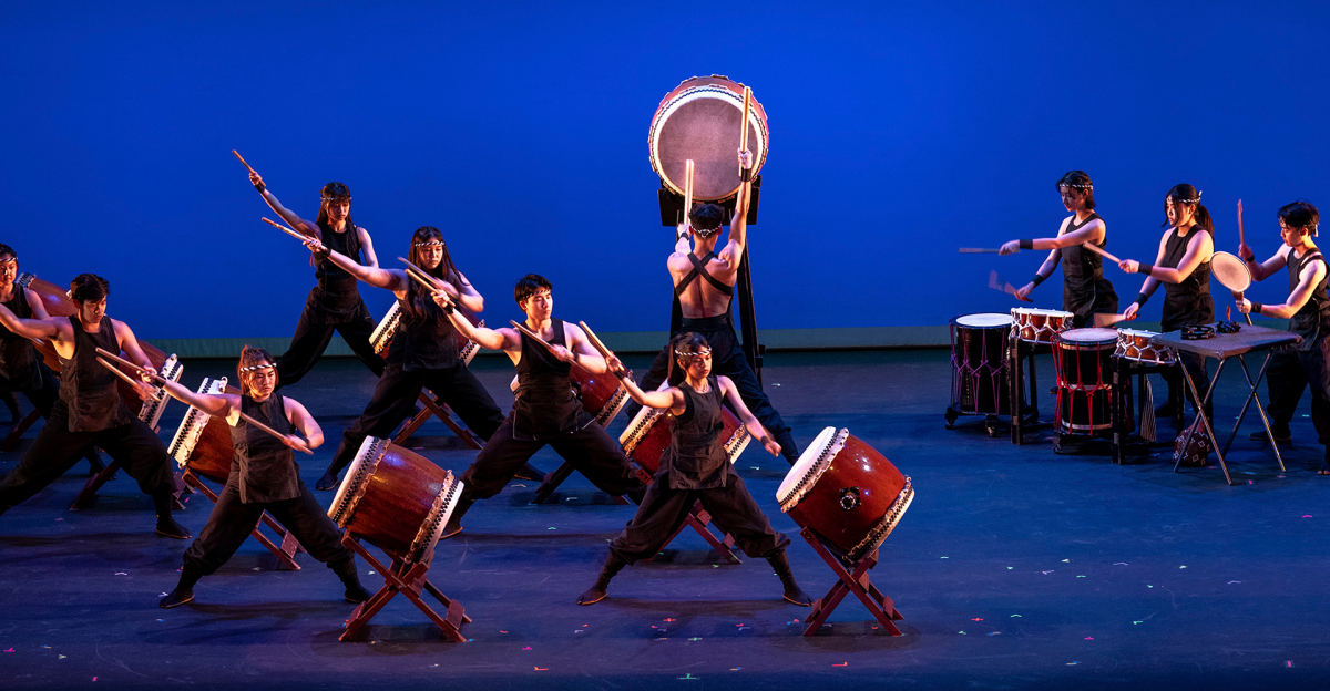 Group of performers playing large wooden taiko drums with leather drumheads on stage