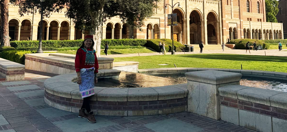 Person in colorful attire by stone fountain, brick building in background.