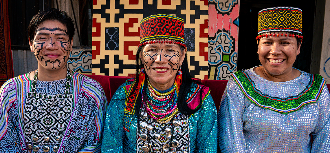 Three people in traditional beaded clothing and hats with facial paint, colorful fabric and silver ornaments