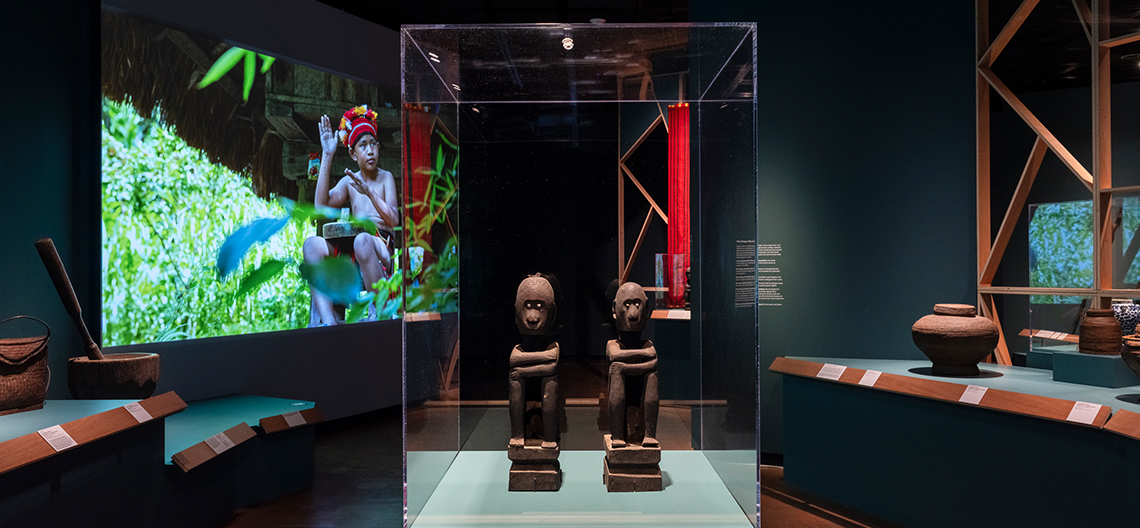 Two wooden tribal statues displayed in a glass case inside a dimly lit museum with teal walls