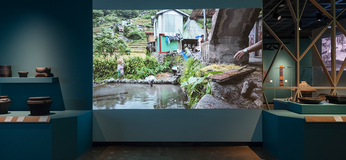 Museum exhibit with ceramic pots displayed on blue shelves and a large photo of a rural scene on the wall